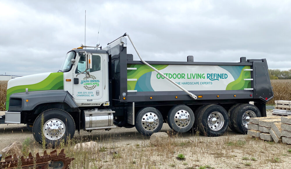 A large black and white dump truck with six wheels is parked on gravel near stacks of stone. The truck&rsquo;s side advertises &ldquo;Outdoor Living Refined&rdquo; and displays a landscaping company&rsquo;s contact information.