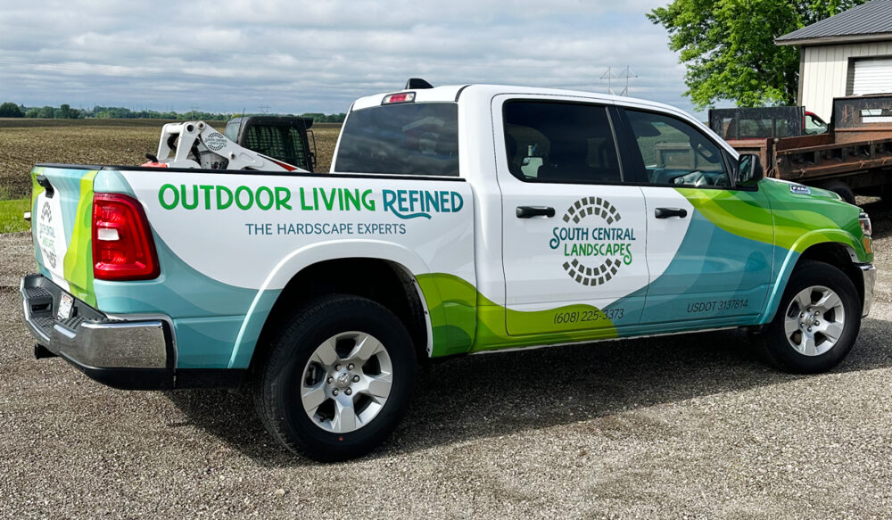 A white pickup truck with green and blue graphics advertising South Central Landscapes, featuring the slogan Outdoor Living Refined: The Hardscape Experts, is parked on a gravel driveway near a field and a building.
