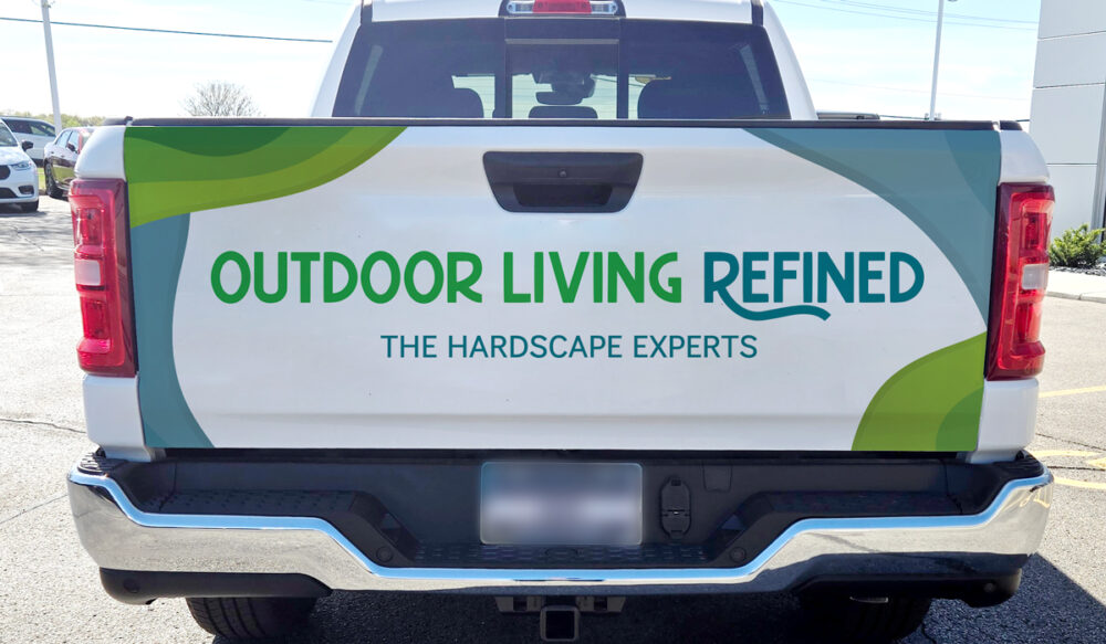 A white pickup truck with a graphic on the tailgate reading OUTDOOR LIVING REFINED and THE HARDSCAPE EXPERTS in green and blue letters, with green and blue abstract shapes in the background.