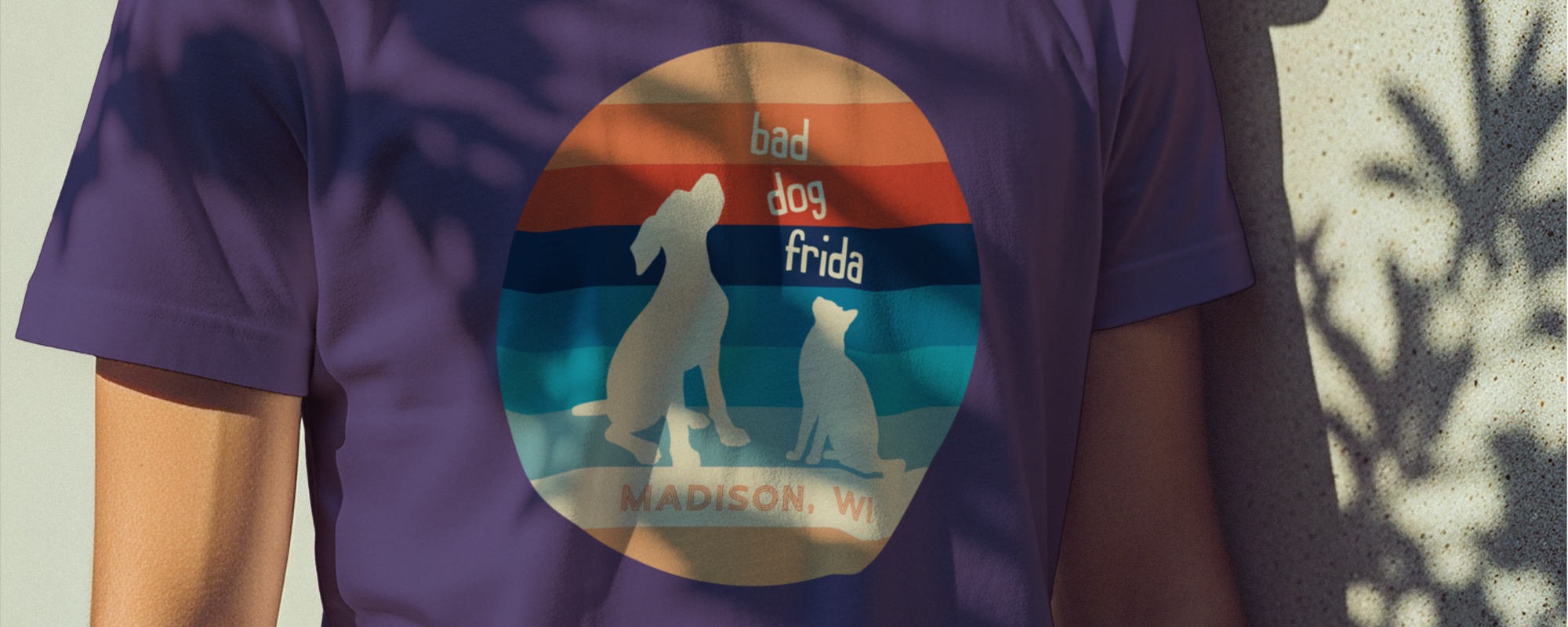 A person wearing a purple t-shirt with a retro-style circular design featuring silhouettes of a dog and a cat, the words bad dog frida, and MADISON, WI below on striped background.
