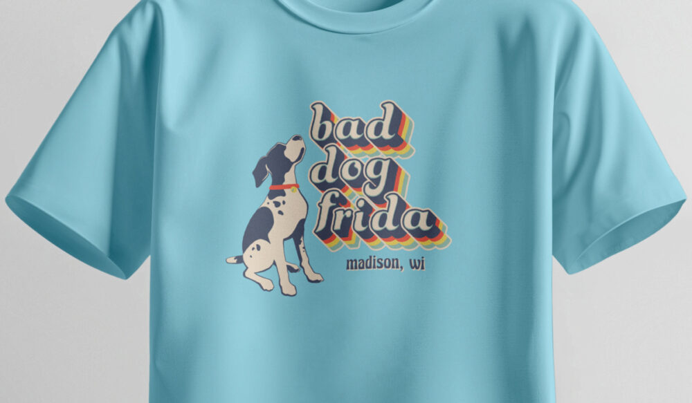 Light blue t-shirt featuring a retro-style graphic of a black and white dog sitting, with the words bad dog frida in colorful, layered text above &ldquo;madison, wi.&rdquo;.