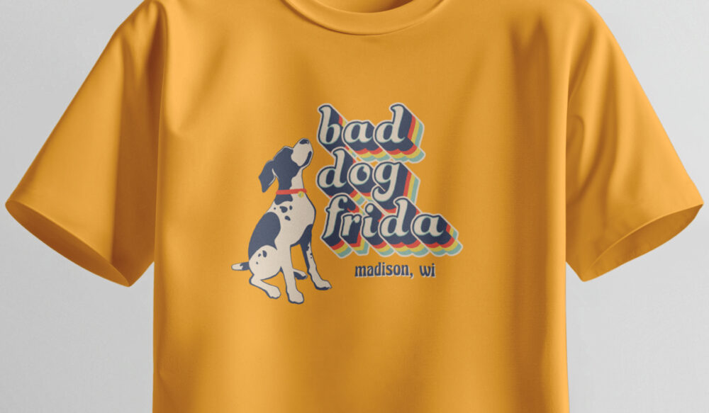 A mustard yellow t-shirt featuring a cartoon dog and the retro-style text bad dog frida in colorful letters, with madison, wi written below.