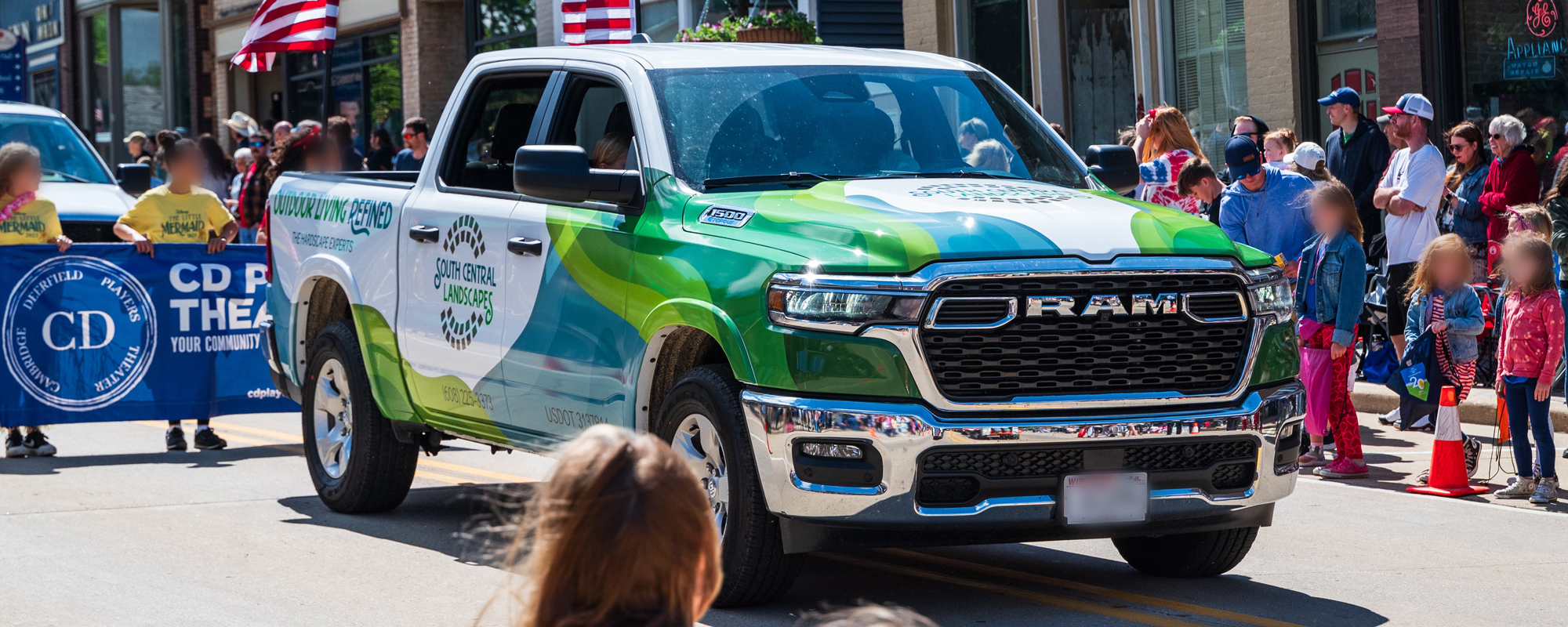 A green and white RAM pickup truck drives in a parade, surrounded by people and children holding a blue banner. American flags are displayed, and spectators watch from the sidewalk on a sunny day.