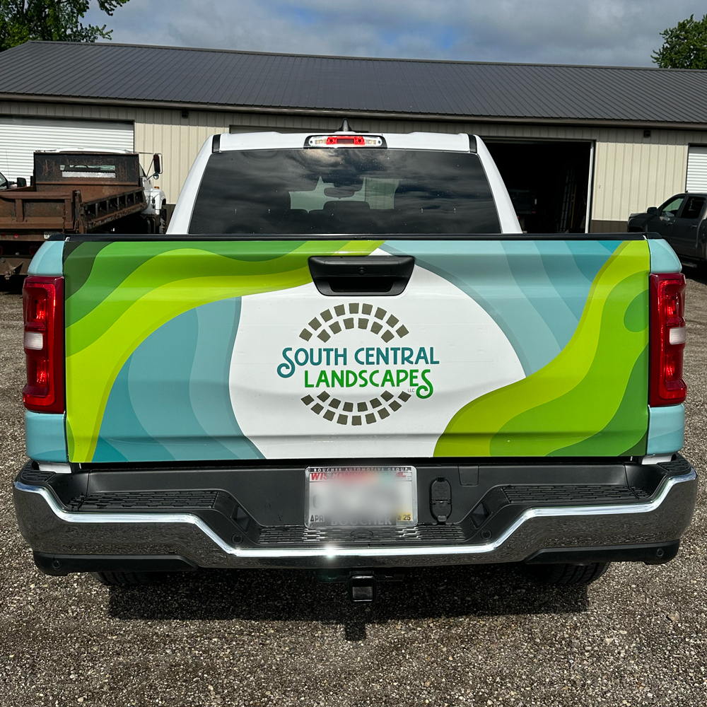 The back of a white pickup truck displays a colorful South Central Landscapes logo with green and blue wave designs on the tailgate, parked outside a tan metal building.
