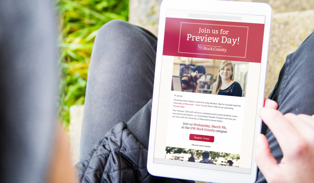 Person holding a tablet displaying an invitation for a Preview Day by UW-Rock County. The screen shows a photo of a woman and event details, encouraging registration. The person is seated outdoors, with grass visible.